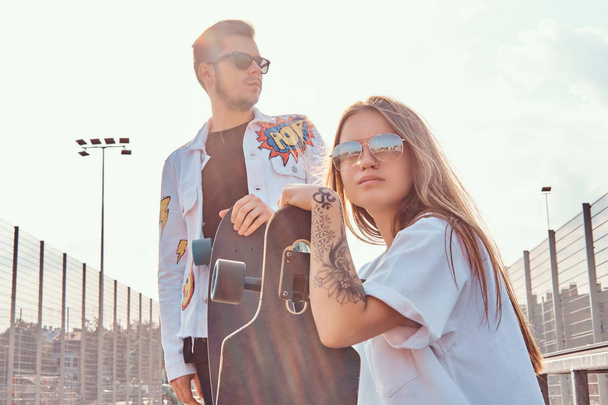 Accogliente coppia di giovani hipster vestiti alla moda con skateboard presso il complesso sportivo cittadino nella giornata di sole, con toni caldi
. - Foto, immagini