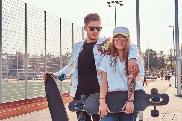 Atractive δυο μοντέρνα ντυμένος νέους οι Χίπστερς ποζάρει με skateboards στην πόλη αθλητικό συγκρότημα ηλιόλουστη ημέρα - Φωτογραφία, εικόνα