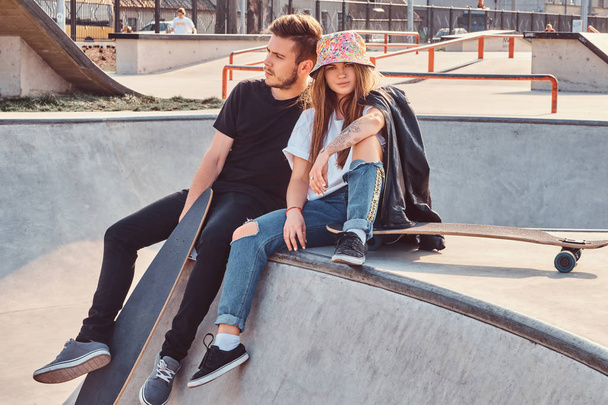 Young hipster couple trendy dressed relaxing together with skateboards on a skatepark on a sunny day. - Photo, Image