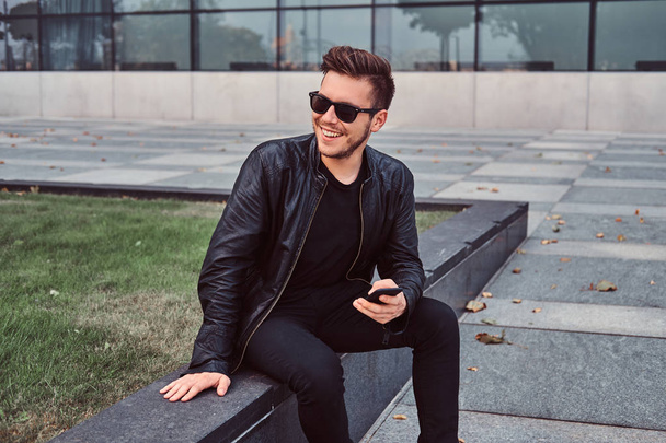Jeune homme souriant avec des lunettes de soleil aux cheveux élégants habillé d'une veste en cuir noir tient smartphone tout en étant assis près d'un gratte-ciel
. - Photo, image