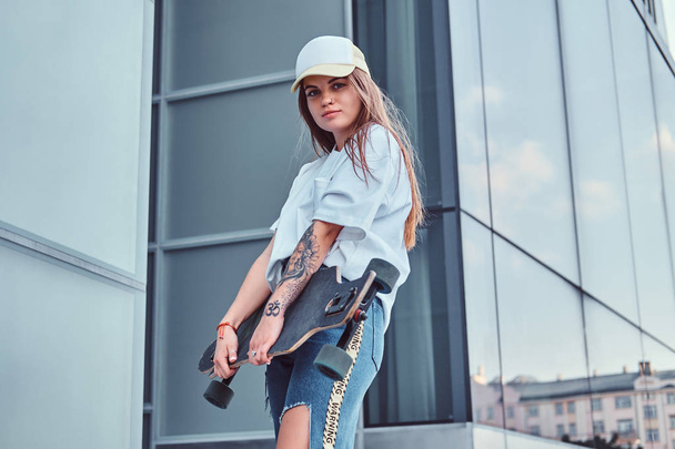 Mladí bokovky dívka v čepici oblečený v bílé košili a roztrhaný džíny drží skateboardu při pózování poblíž mrakodrap. - Fotografie, Obrázek