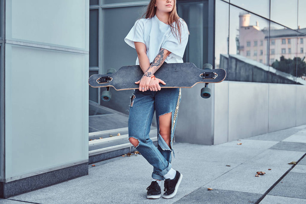 Cropped photo of a young hipster girl dressed in white shirt and ripped jeans holds skateboard while posing near skyscraper. - Photo, Image