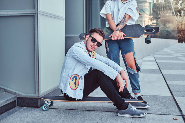 Trendy dressed couple - young hipster man sitting on a longboard and his girlfriend standing near outdoors. - Foto, afbeelding