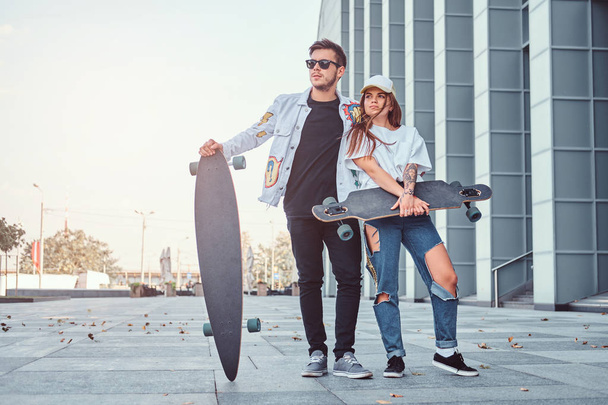 Jeune couple attrayant habillé de vêtements à la mode posant avec des planches à roulettes près du gratte-ciel
. - Photo, image