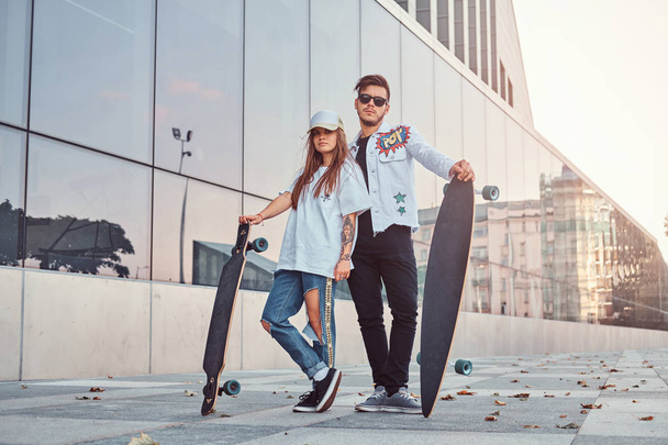 Attractive young couple dressed in trendy clothes posing with skateboards near skyscraper. - Zdjęcie, obraz
