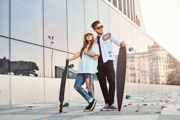 Attraente giovane coppia vestita con abiti alla moda in posa con skateboard vicino al grattacielo
. - Foto, immagini