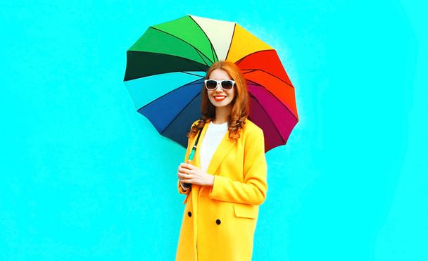 Mode glücklich lächelnde Frau mit buntem Regenschirm in gelber Jacke auf blauem Hintergrund - Foto, Bild