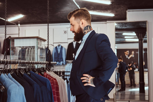 Elegantemente vestido hombre barbudo con tatuajes en las manos y el cuello elige nuevo traje en la tienda de ropa de hombre
. - Foto, imagen