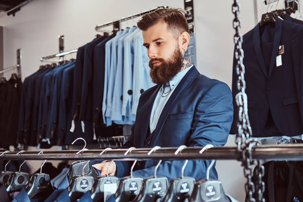 Элегантно одетый бородатый мужчина с татуировками на руках и шее выбирает новый костюм в магазине мужской одежды
. - Фото, изображение