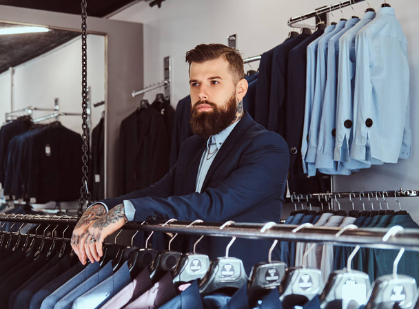 Nadenkend tattoed mannelijke met stijlvolle baard en haren gekleed in elegante pak staande in een herenkleding winkel. - Foto, afbeelding