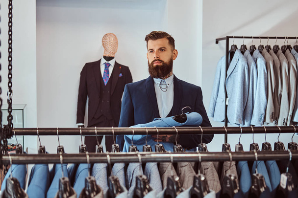 Elegantemente vestido hombre barbudo con tatuajes en las manos y el cuello elige nuevo traje en la tienda de ropa de hombre
. - Foto, imagen