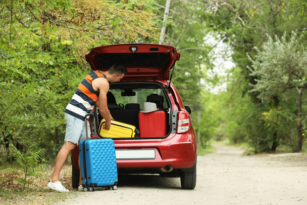 Happy man packing suitcases into car trunk near forest. Space for text - Фото, изображение
