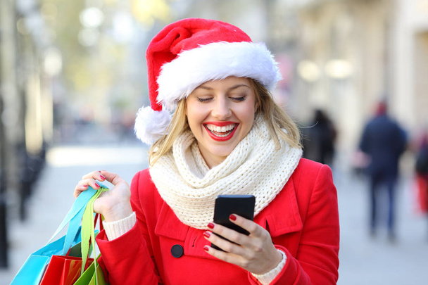 Front view portrait of a happy shopper holding shopping bags checking phone on christmas in the street - Fotografie, Obrázek