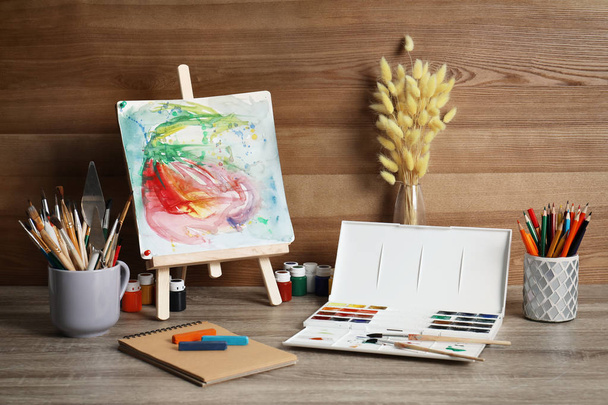 Easel with abstract painting and set of professional art supplies on table against wooden background - Foto, afbeelding
