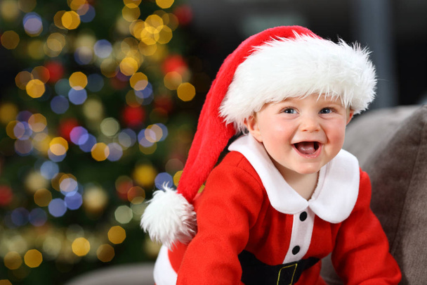 Baby laughing wearing santa claus disguise on a couch at home in christmas with a tree in the background - Фото, зображення