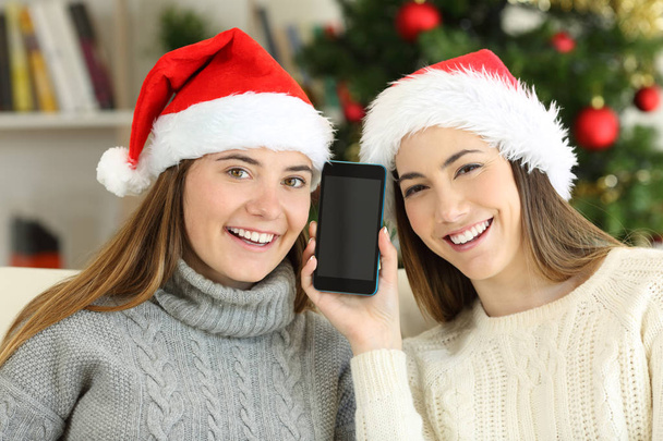 glückliche Mädchen mit leerem Handy-Bildschirm an Weihnachten, die zu Hause auf einer Couch im Wohnzimmer sitzen - Foto, Bild