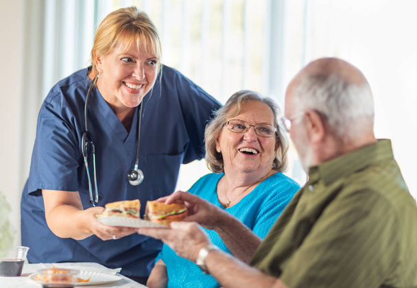 Female Doctor or Nurse Serving Senior Adult Couple Sandwiches at Table. - Фото, изображение