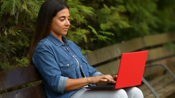 Happy woman chating online with a laptop and laughing sitting on a bench in a park - Filmmaterial, Video
