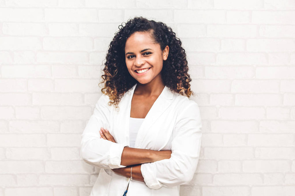 Sorridente bella business professionale afro-americana donna nera con le braccia incrociate in piedi vicino a sfondo muro di mattoni bianchi
 - Foto, immagini