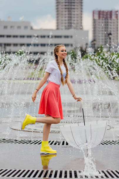 Young pretty girl with two braids in yellow boots and with transparent umbrella stands near fountain. - Foto, afbeelding