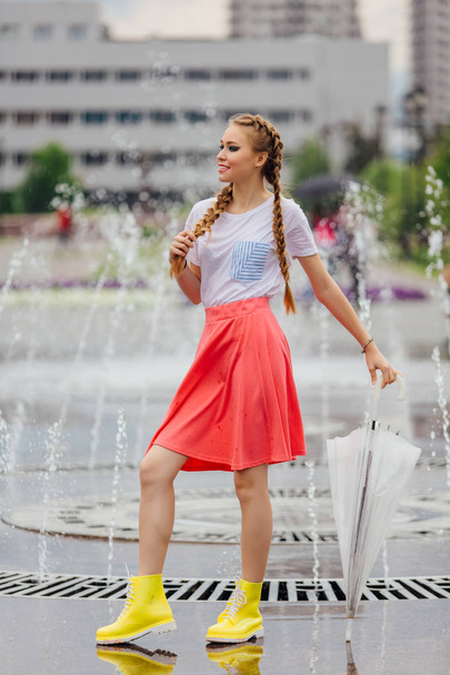 Young pretty girl with two braids in yellow boots and with transparent umbrella stands near fountain. - Photo, image