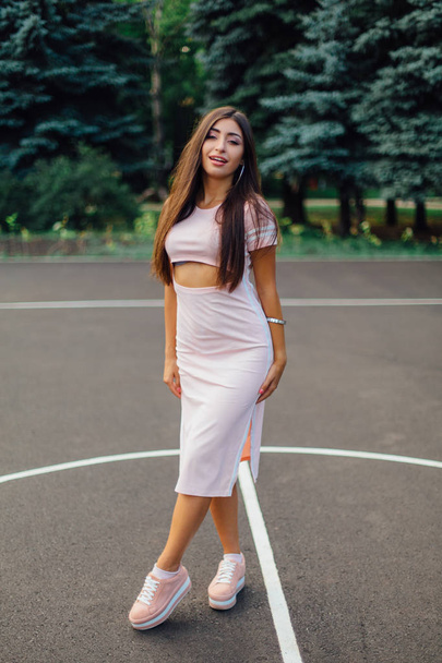 Charming brunette female dressed in a pink dress posing on the basketball court during sunset. - Zdjęcie, obraz