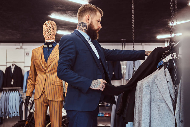 Homme barbu élégamment habillé avec des tatouages sur les mains et le cou choisit un nouveau costume dans un magasin de vêtements pour hommes
. - Photo, image