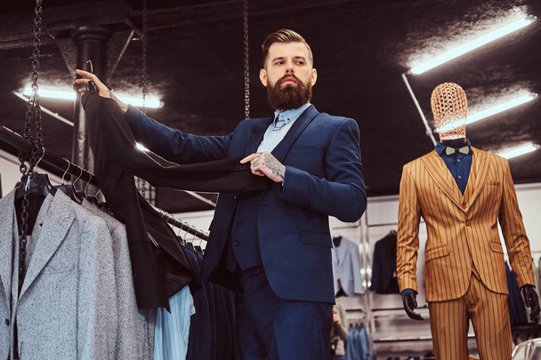 elegant gekleideter bärtiger Mann mit Tätowierungen an Händen und Hals wählt neuen Anzug in einem Herrengeschäft. - Foto, Bild