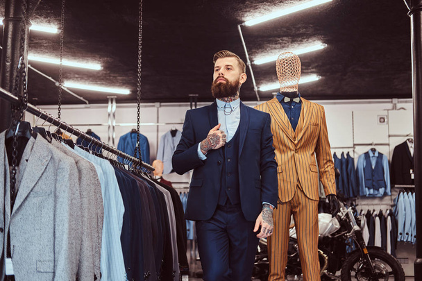 Assistant de magasin barbu élégamment habillé avec debout près du mannequin dans un magasin de vêtements pour hommes
. - Photo, image