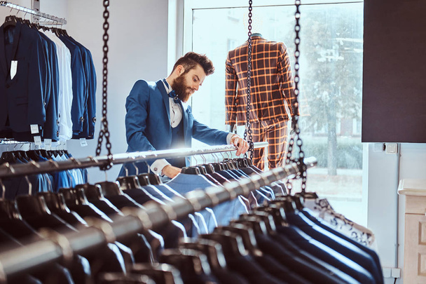 Бородатый помощник магазина, одетый в синий элегантный костюм, работает в магазине мужской одежды
. - Фото, изображение
