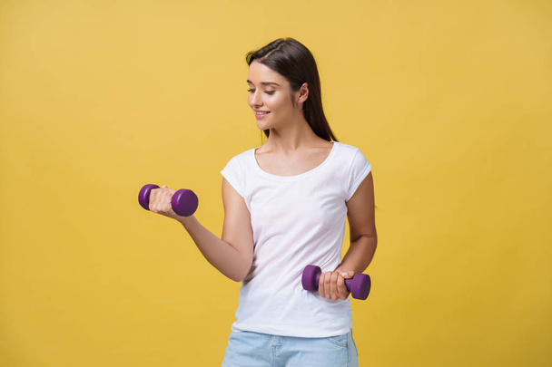 Aufnahme einer schönen und sportlichen jungen Frau, die vor gelbem Hintergrund Gewichte stemmt. - Foto, Bild