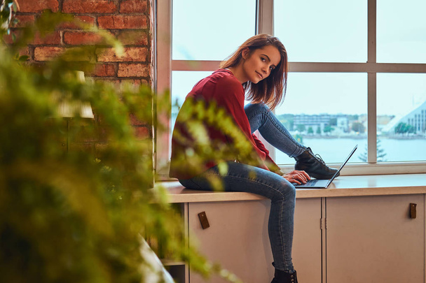 junge schöne Studentin sitzt mit Laptop auf Fensterbank im Studentenwohnheim. - Foto, Bild