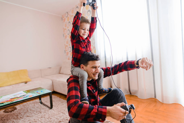 alegre padre emocionado y pequeño hijo en camisas rojas jugando juego de consola con gamepads
 - Foto, Imagen