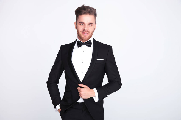 portrait of relaxed stylish man buttoning his black suit while standing on light grey background - Photo, Image
