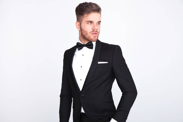 portrait of stylish curious man wearing black tuxeo and bowtie looking to side while standing on light grey background - Фото, изображение