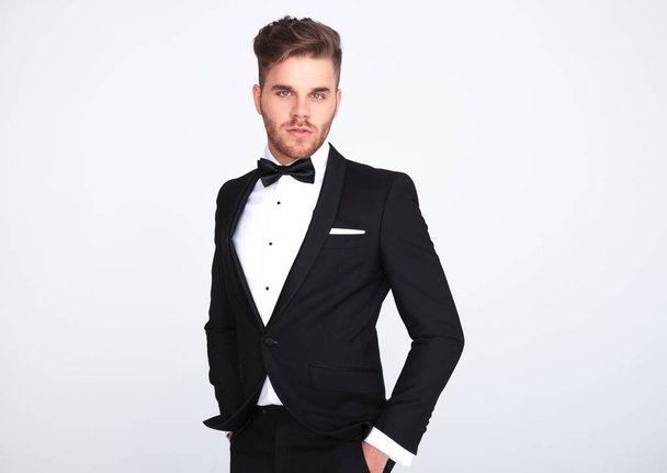 portrait of relaxed elegant man in black tuxedo standing on light grey background with hands in pockets - Photo, Image