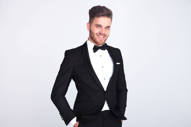 portrait of relaxed smiling gentleman in black tuxedo standing on light grey background - Photo, Image