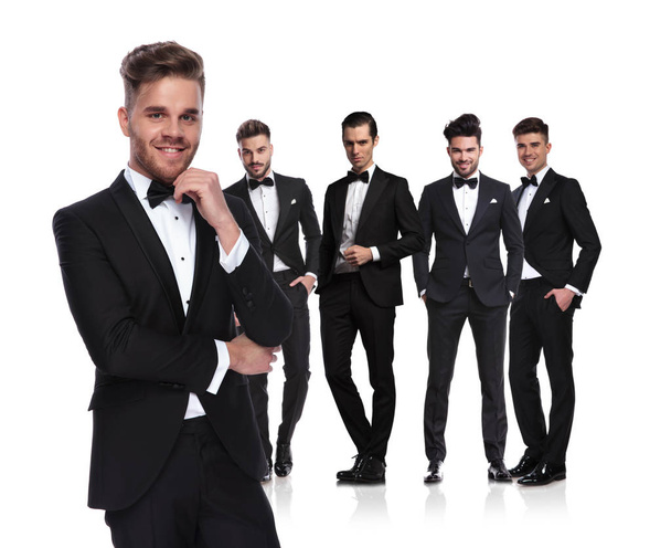 young group leader in tuxedo stands in front of his team on white background and smiles, looking pensive - Photo, Image