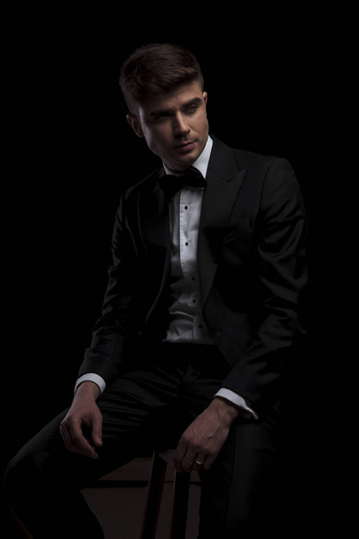 seated stylish man in black tuxedo looks to side on black background - Фото, изображение