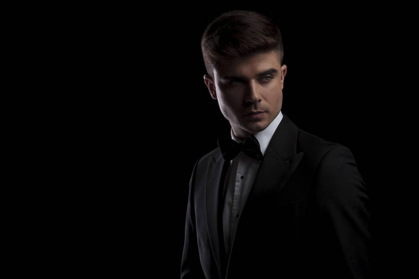 portrait of handsome young man in black tuxedo looking to side while standing on black background - Foto, afbeelding