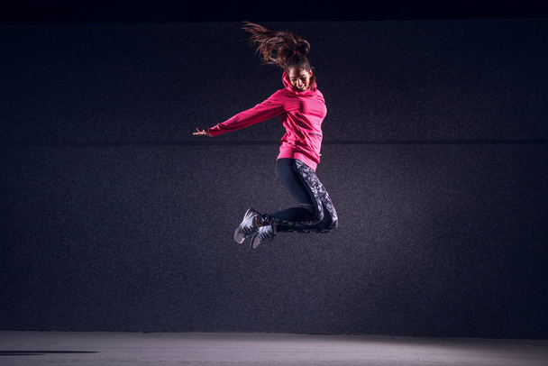 portrait of young slim woman in sportswear jumping against dark wall - Fotoğraf, Görsel