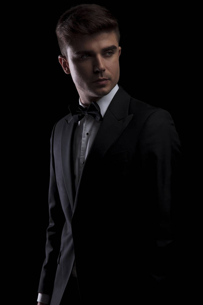 retrato de hombre curioso en esmoquin negro mirando a un lado mientras está de pie sobre fondo negro
 - Foto, Imagen