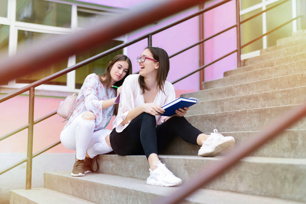 две улыбающиеся симпатичные старшеклассницы читают заметки вместе на лестнице и веселятся
 - Фото, изображение