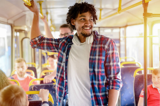 junger fröhlicher Afroamerikaner, der mit Kopfhörern um den Hals in einem Bus nach einer Bar greift. lächelnd und wegschauend. - Foto, Bild
