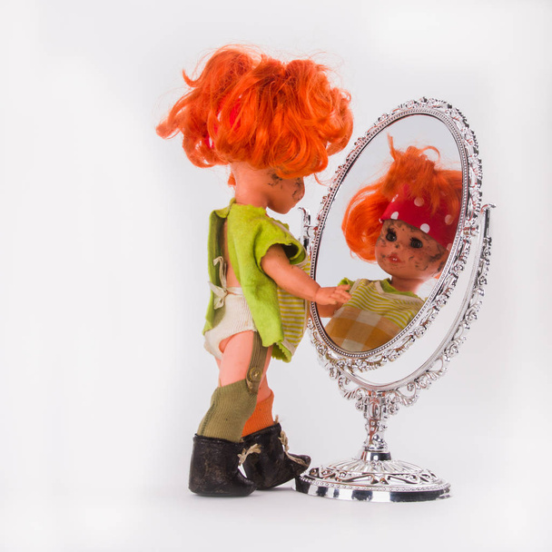 Ακατάστατος μικρό κορίτσι βλέποντας τον εαυτό της. Κούκλα με τα κόκκινα μαλλιά κοιτάζει τον εαυτό της στον καθρέφτη και να παίζει με αντανάκλαση. - Φωτογραφία, εικόνα