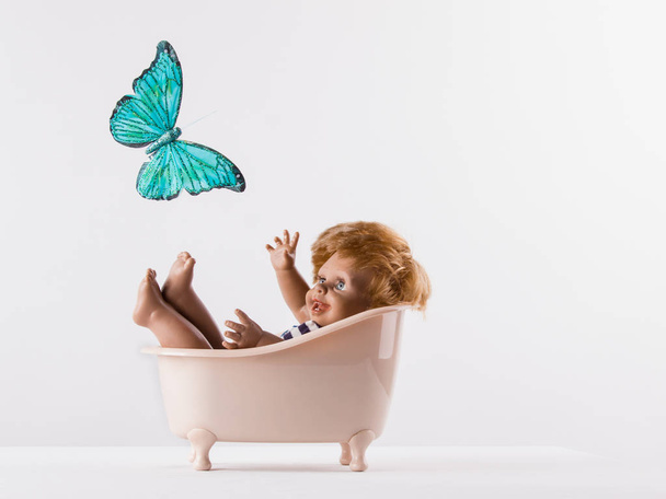 A small doll-boy lies in the bathroom on a white background and looking at a beautiful flying butterfly - Foto, Imagem