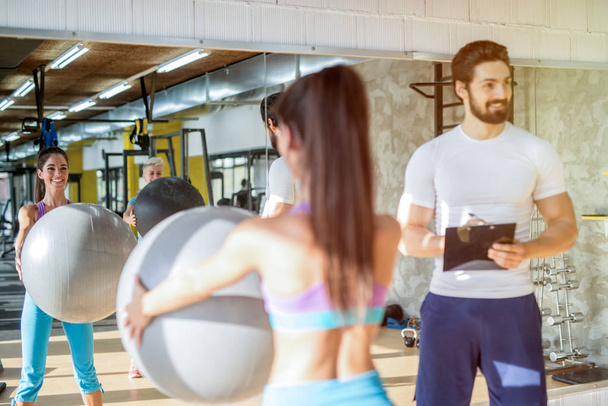 Picture of personal fitness trainer talking with his female client while she is holding big ball in her hands. - Photo, Image