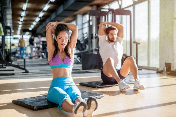 Bild eines gut aussehenden sportlichen Paares, das in einem hellen Fitnessstudio sitzt und nach dem Training seine Armmuskeln dehnt. - Foto, Bild