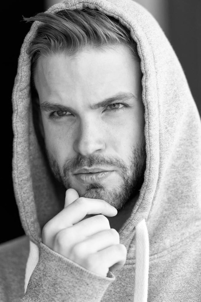 Guy bearded and attractive touches bristle on chin. Masculinity concept. Man with beard or unshaven guy looks handsome hooded. Man with bristle on serious face, urban background, defocused - Fotó, kép