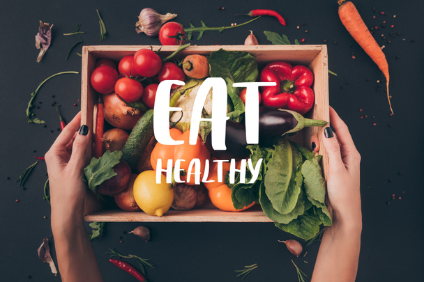 cropped image of woman holding wooden box with vegetables above gray table with "eat healthy" lettering - Photo, Image